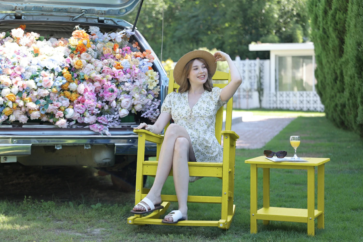 Woman sitting in a yellow chair next to a car filled with flowers, with a table and sunglasses nearby.