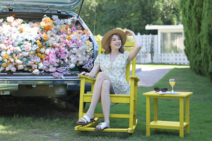 Woman sitting in a yellow chair next to a car filled with flowers, with a table and sunglasses nearby.