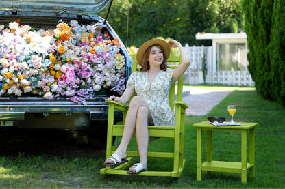 A woman in a floral dress and straw hat relaxes in an apple green wooden rocking chair beside an open car trunk filled with colorful artificial flowers, with a matching green side table holding sunglasses and an orange drink on a grassy lawn.