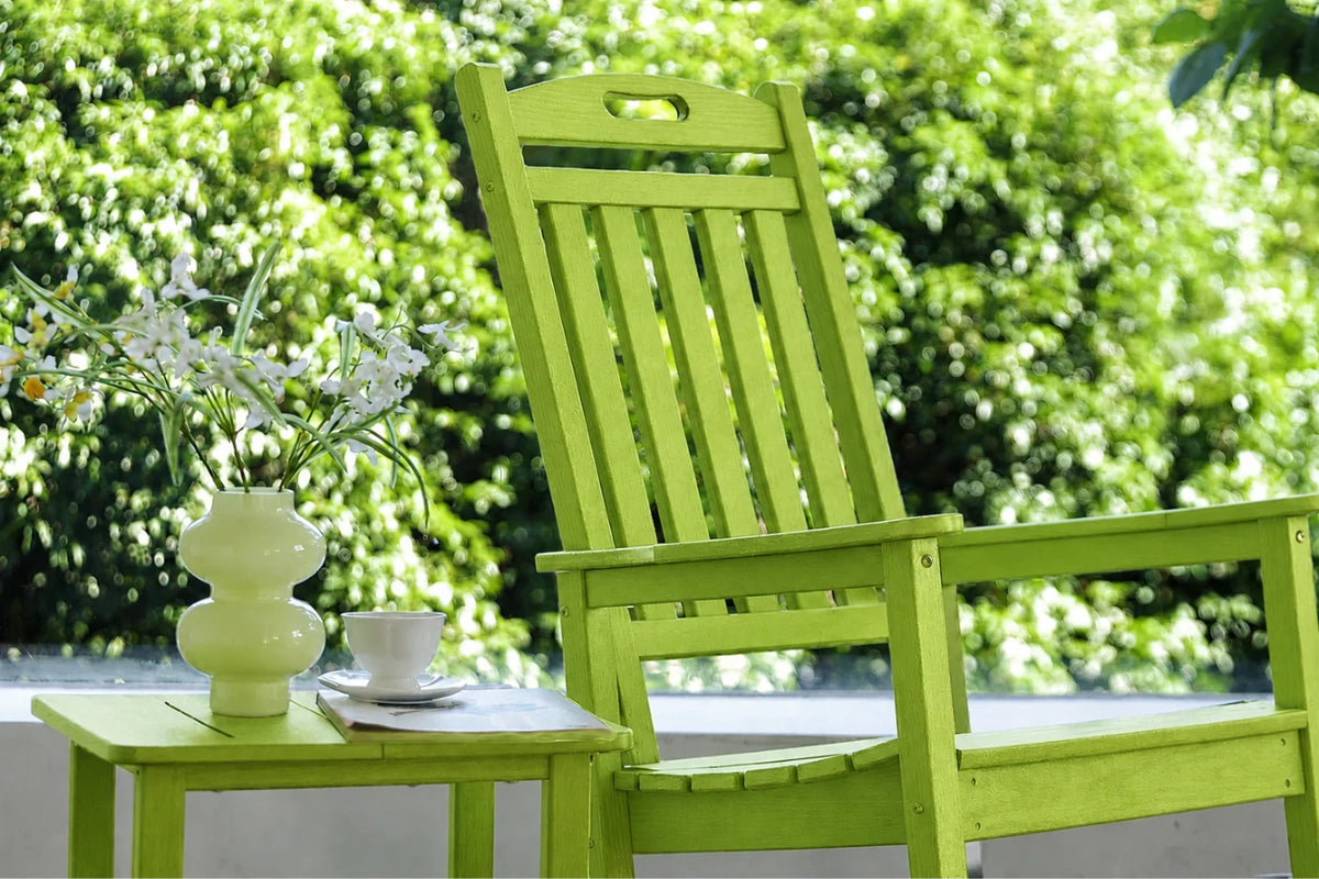 An apple green outdoor wooden chair with a matching side table holding a white vase of flowers and a white cup, set against a blurred green garden background.