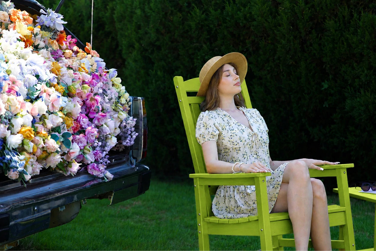 A woman wearing a straw hat and a cream floral dress sits in an apple green outdoor chair next to a black truck bed overflowing with colorful artificial flowers, set against a dark green hedge background.