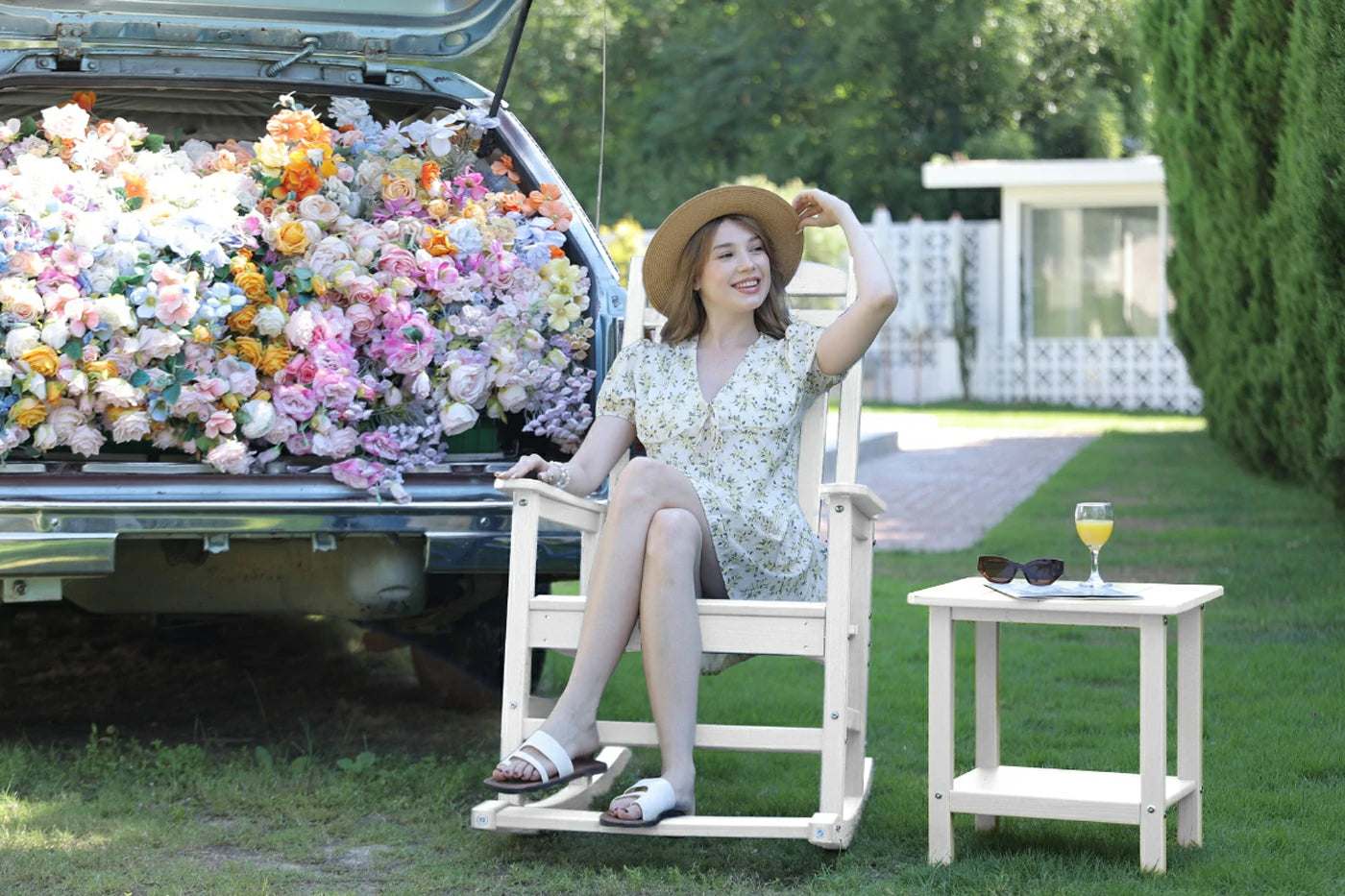 Woman sitting on a white chair next to a car filled with flowers, with a small table and sunglasses in the background.
