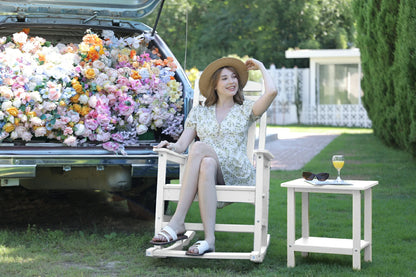 Woman sitting on a white chair next to a car filled with flowers, with a small table and sunglasses in the background.
