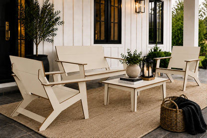 A coordinated outdoor patio furniture set on a porch, including two beige Adirondack-style chairs, a matching loveseat, a small coffee table, and a natural jute rug. Decorative plants, lanterns, books, and a wicker basket with a dark throw are arranged on the porch in front of a white house with black-framed windows.