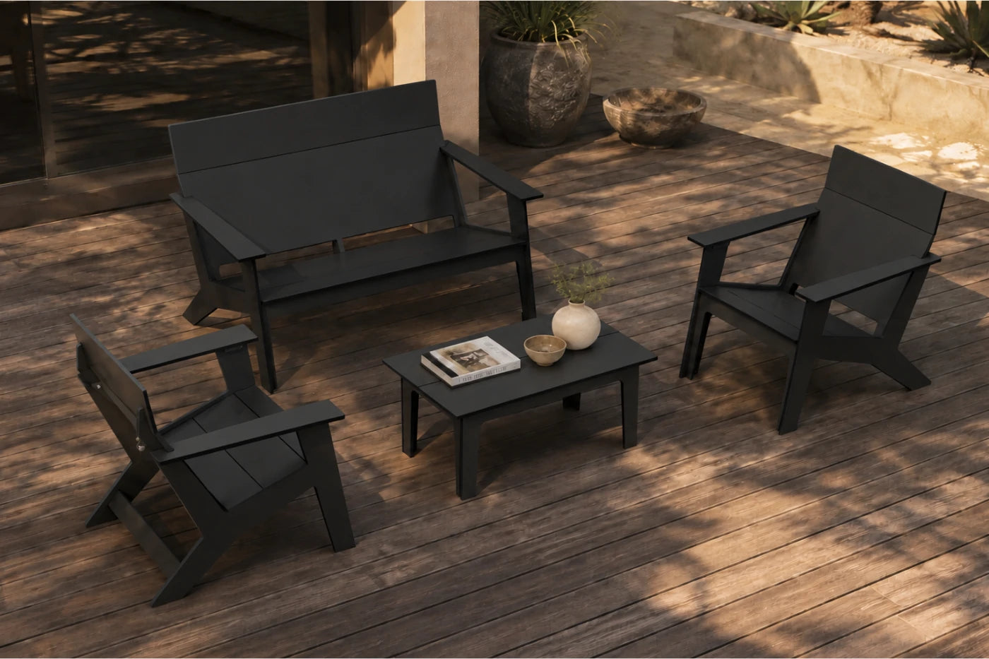 A four-piece outdoor patio furniture set in black, including a loveseat, two armchairs, and a small rectangular coffee table, arranged on a wooden deck with decorative props.