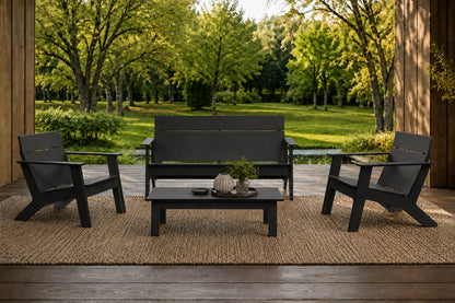 A 4-piece outdoor patio furniture set including a loveseat, two armchairs, and a coffee table, all in a black finish, arranged on a woven jute rug on a wooden porch with a lush green garden background.