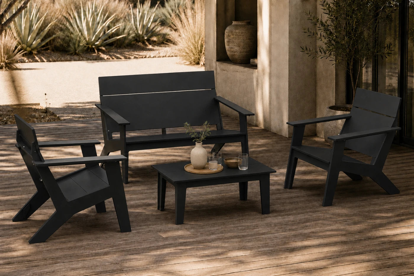 A four-piece outdoor patio furniture set in matte black, including a loveseat, two armchairs, and a small square coffee table, arranged on a wooden deck with plants and a stucco wall in the background.