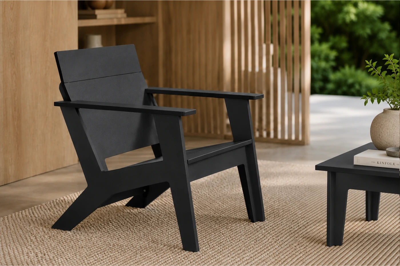 A modern black outdoor armchair with a wide armrest and angled legs, shown in a styled patio setting with a small matching side table, woven rug, wooden slat screen, and greenery in the background.