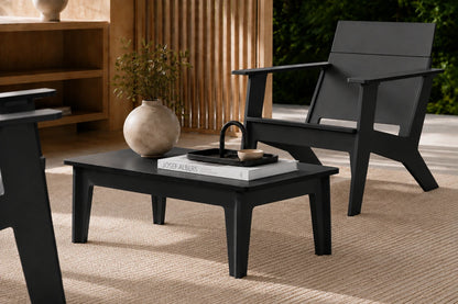 A modern black outdoor coffee table with a matching black chair in a styled patio setting, featuring a woven rug, wooden slat screen, and a ceramic vase on the table.