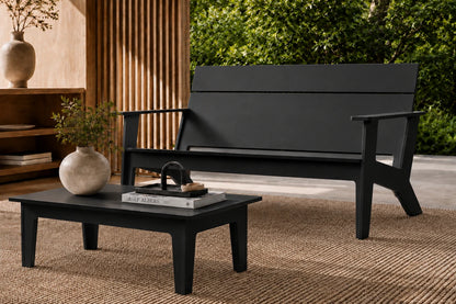 A modern black outdoor loveseat with a matching rectangular coffee table, styled on a woven jute rug in a patio setting with a wooden slatted screen and green foliage in the background.