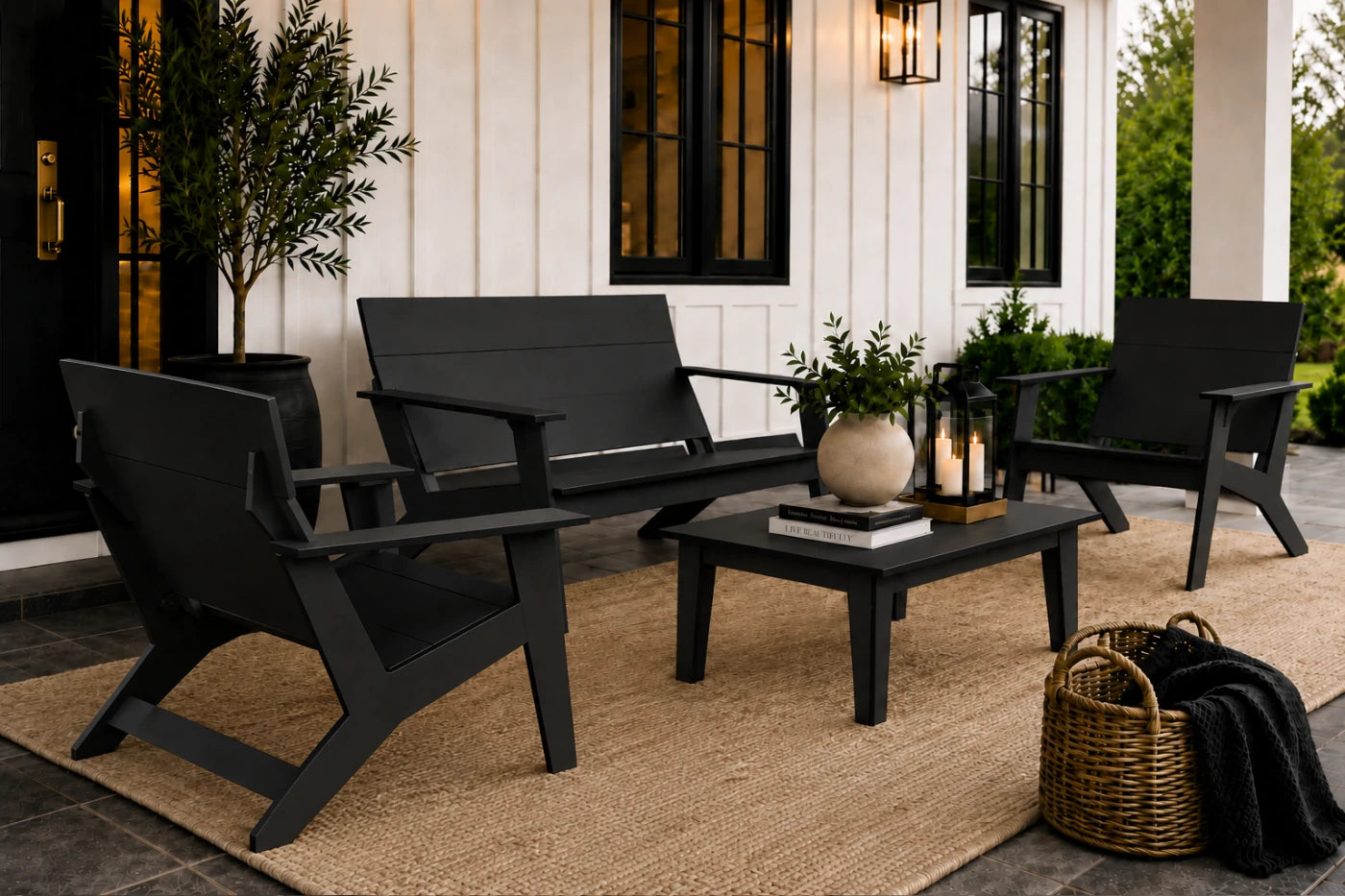 A set of four black outdoor patio furniture pieces arranged on a jute rug in front of a white house with black-framed windows. The set includes a loveseat, two armchairs, and a small rectangular coffee table with decorative items on top.