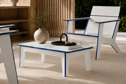 A modern white outdoor chair with blue trim and a matching white coffee table in a sunlit patio setting. The table is styled with a beige ceramic vase, a black metal tea infuser, and a book.