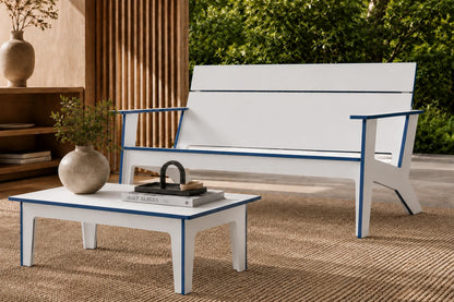 A modern outdoor patio chair and matching coffee table in white with blue trim, styled in a warm, sunlit patio setting with a woven rug, wooden slat wall, and green foliage in the background.
