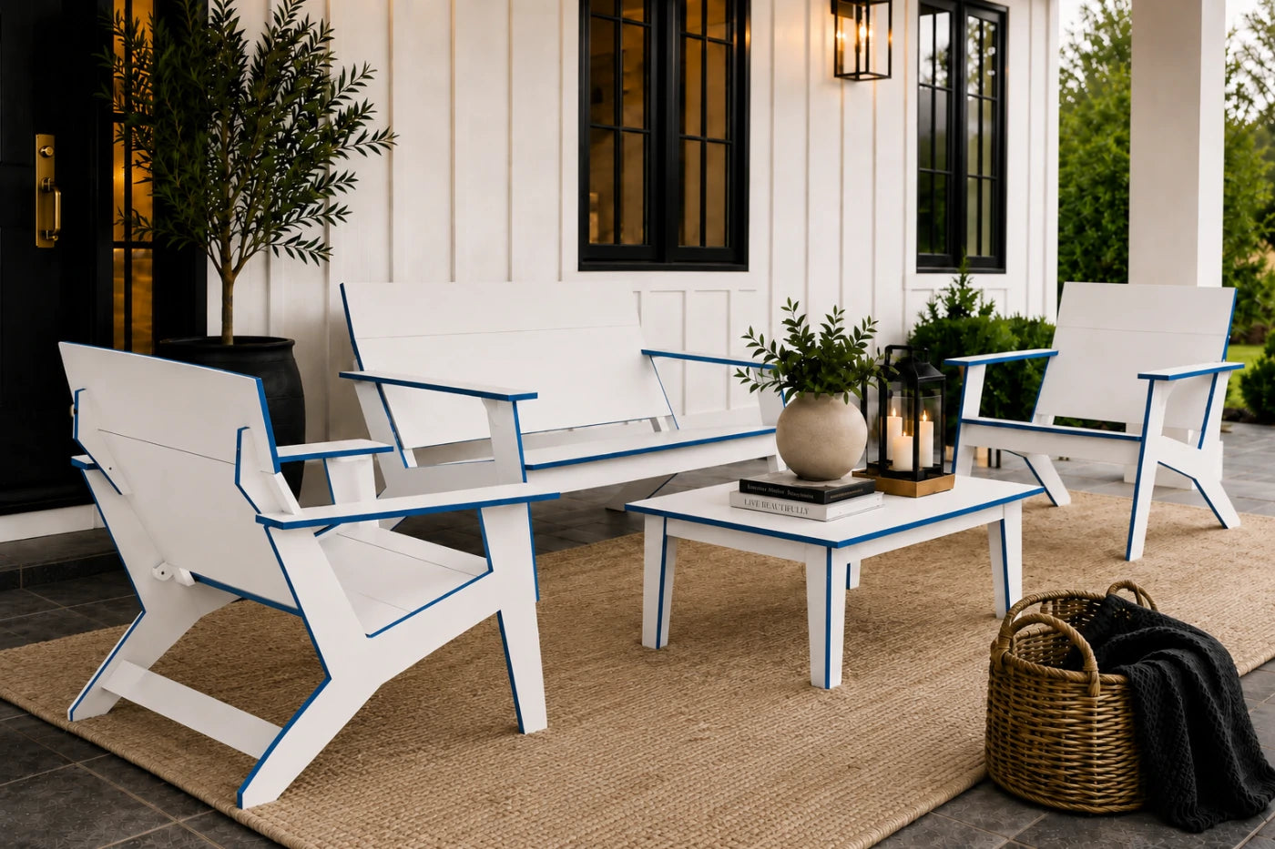 A modern outdoor furniture set on a porch, including a white loveseat with blue trim, two matching armchairs, a small white coffee table, and a beige jute rug. The set is styled with plants, a lantern, and a wicker basket on a gray stone patio in front of a white house with black-trimmed windows.