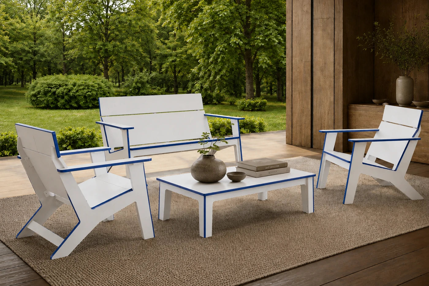 A four-piece outdoor patio furniture set including two Adirondack chairs, a loveseat, and a coffee table, finished in white with blue trim, arranged on a jute rug in a patio setting with a green garden background.