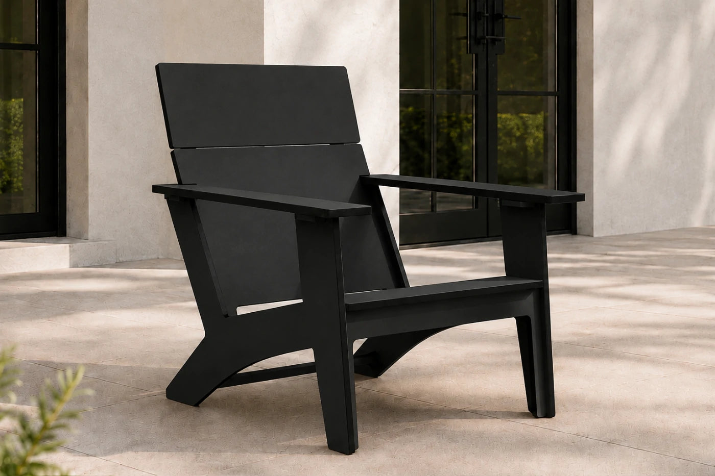 A modern black outdoor Adirondack chair with a wide back and armrests, positioned on a light stone patio in front of a building with dark-framed glass doors.
