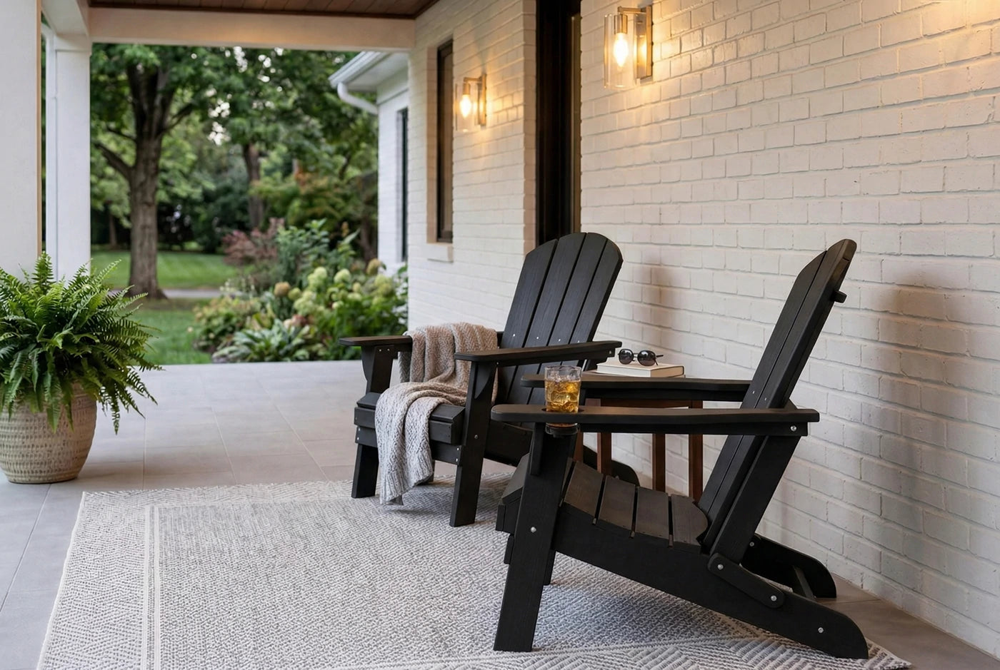 Two black Adirondack chairs on a porch with a white brick wall and outdoor plants.