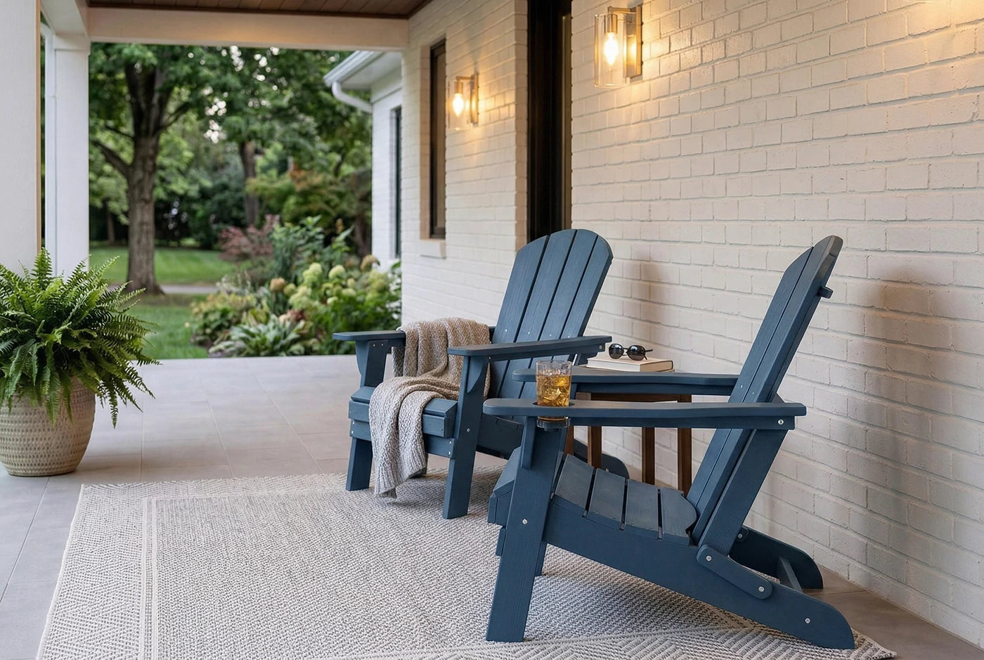 Two blue Adirondack chairs on a porch with a white brick wall and potted plant.