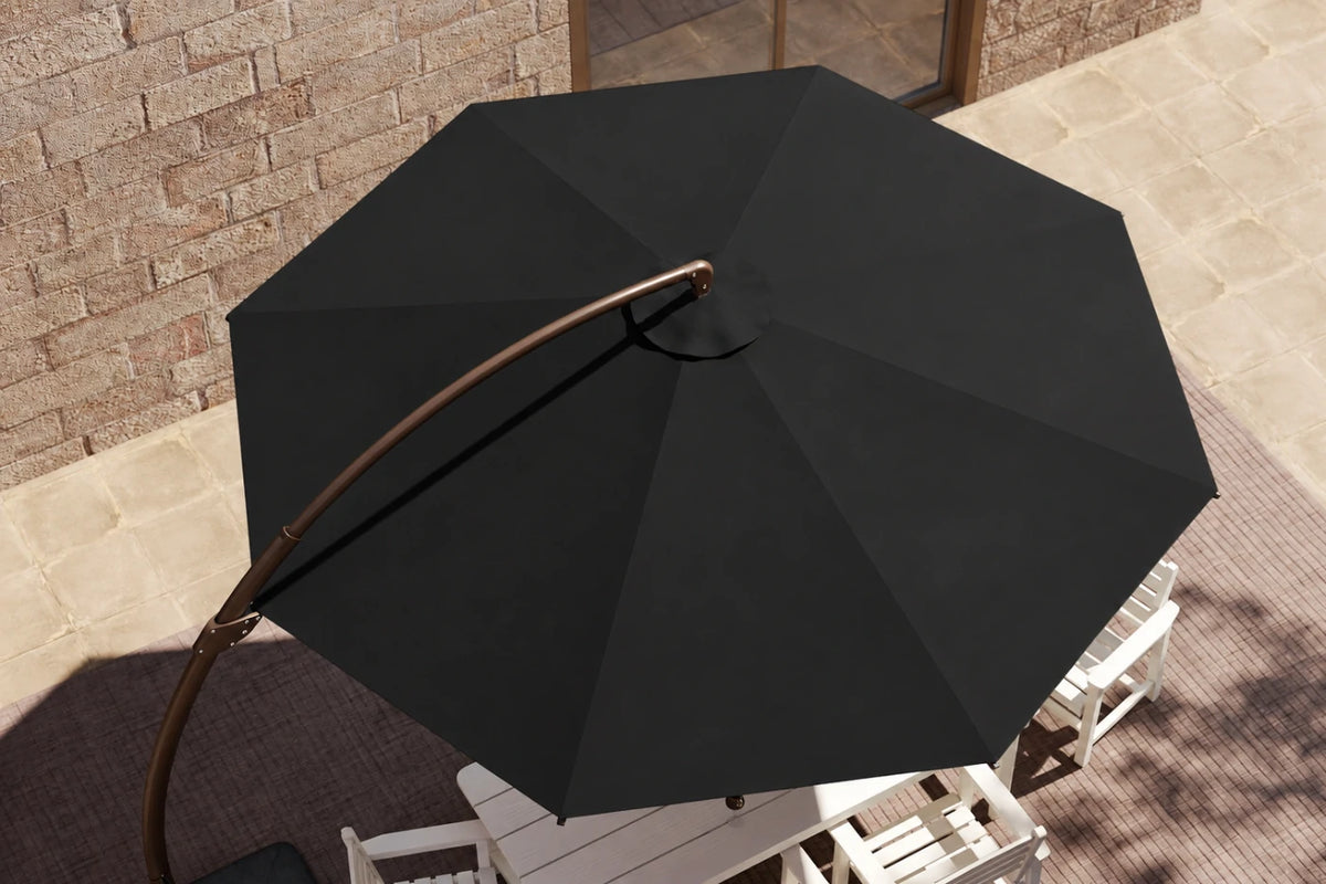 A large black cantilever patio umbrella with a curved bronze-finished metal arm, shown from an elevated angle in an outdoor patio setting with a brick wall and white chairs.