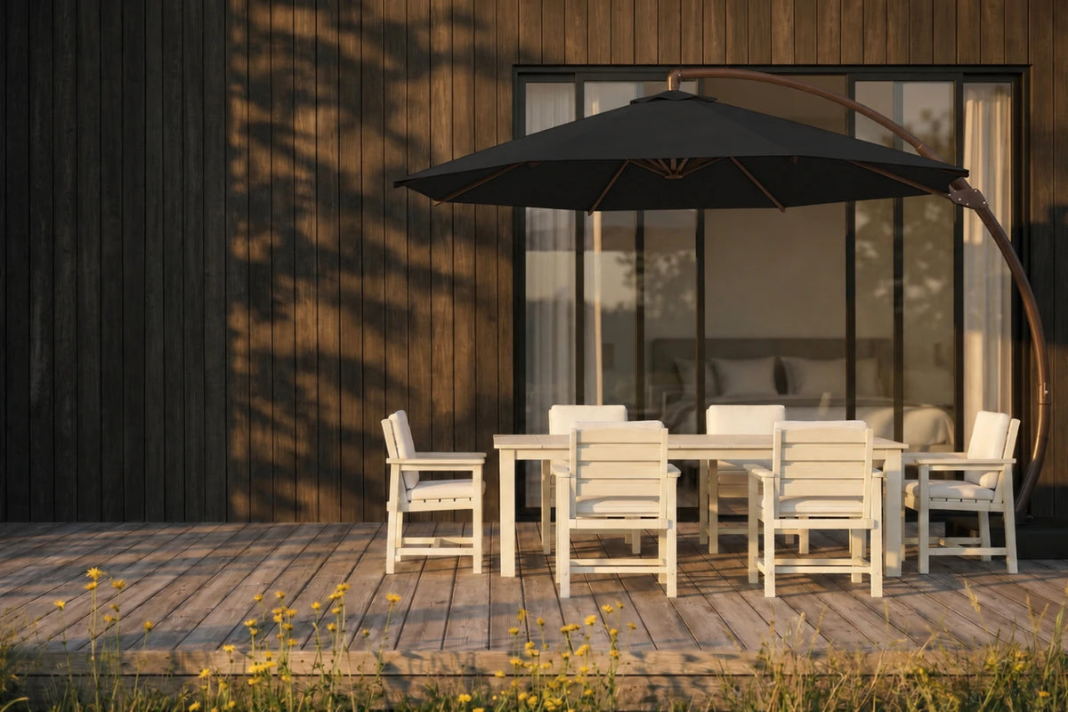 A black patio umbrella with a curved dark metal arm stands over a white outdoor dining table and chairs on a wooden deck in front of a modern house with dark wood siding.