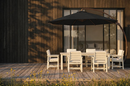 Outdoor dining set on a wooden deck beside a house, featuring a rectangular table, six white chairs, a large black patio umbrella, and a curved black arc umbrella stand.