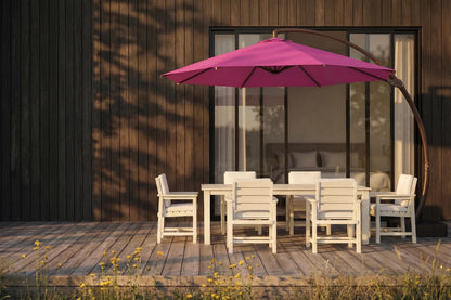 A rose pink patio umbrella with a curved dark metal arm stands over a white outdoor dining table and chairs on a wooden deck in front of a modern house with dark wood siding.