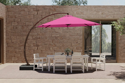 A large outdoor patio setup featuring a bright pink cantilever umbrella over a white dining table and six matching chairs, set against a stone wall background.