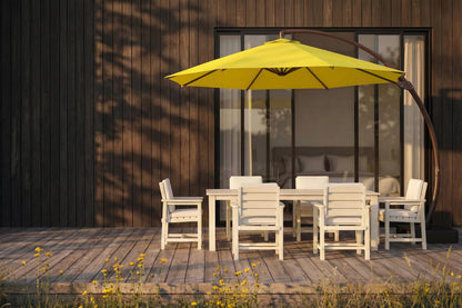 A yellow patio umbrella with a curved dark metal arm stands over a white outdoor dining table and chairs on a wooden deck in front of a modern house with dark wood siding.