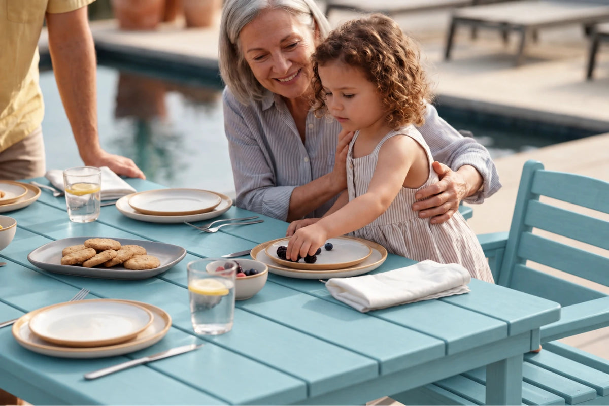 A grandmother and young daughter share a meal at an outdoor patio table by a pool. The table is Aruba blue with plates, glasses, and a plate of cookies.