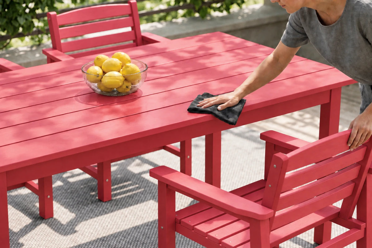 A red outdoor dining table and matching chairs on a patio, with a person wiping the tabletop with a black cloth. A glass bowl of lemons sits on the table.