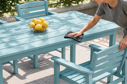 A person wipes down an Aruba blue outdoor dining table with a dark cloth. The table is set with a glass bowl of lemons and surrounded by matching light blue chairs in an outdoor patio setting.