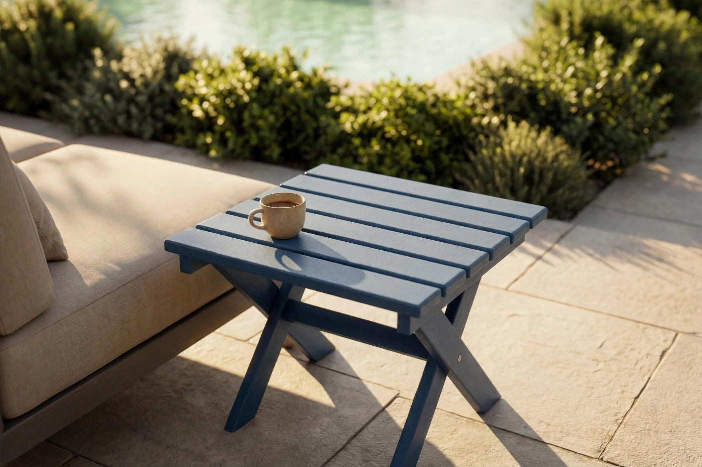 Blue outdoor table with a cup on it, next to a beige sofa by a pool.