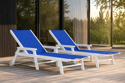 Two modern outdoor chaise lounge chairs with blue fabric cushions and white frames, positioned on a light wood deck in front of a glass-walled building.