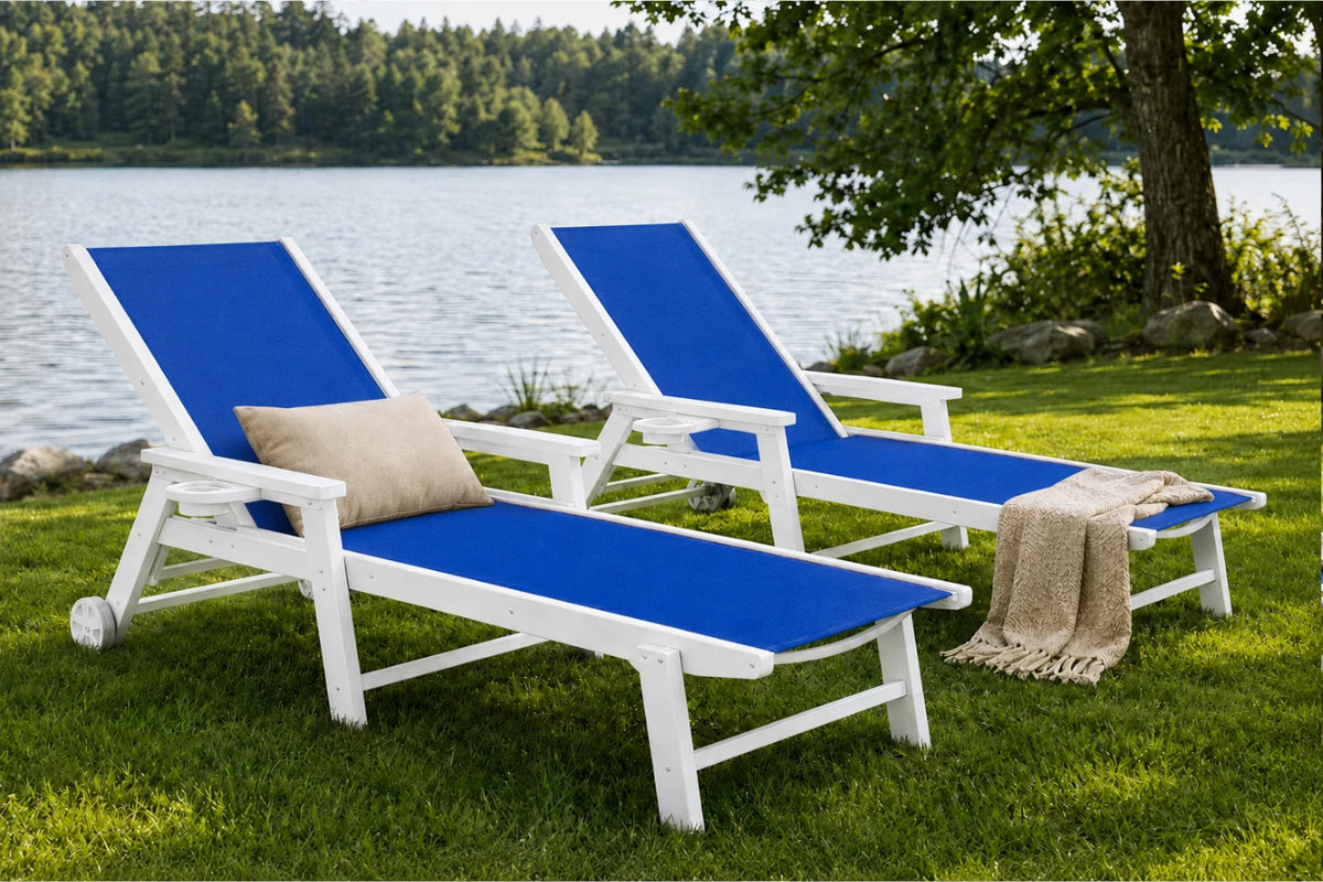 Two outdoor chaise lounge chairs with white frames and blue fabric seats are placed on a grassy lawn beside a lake, with a tree and shoreline in the background. One chair has a beige pillow, and a beige throw blanket is draped over the second chair.