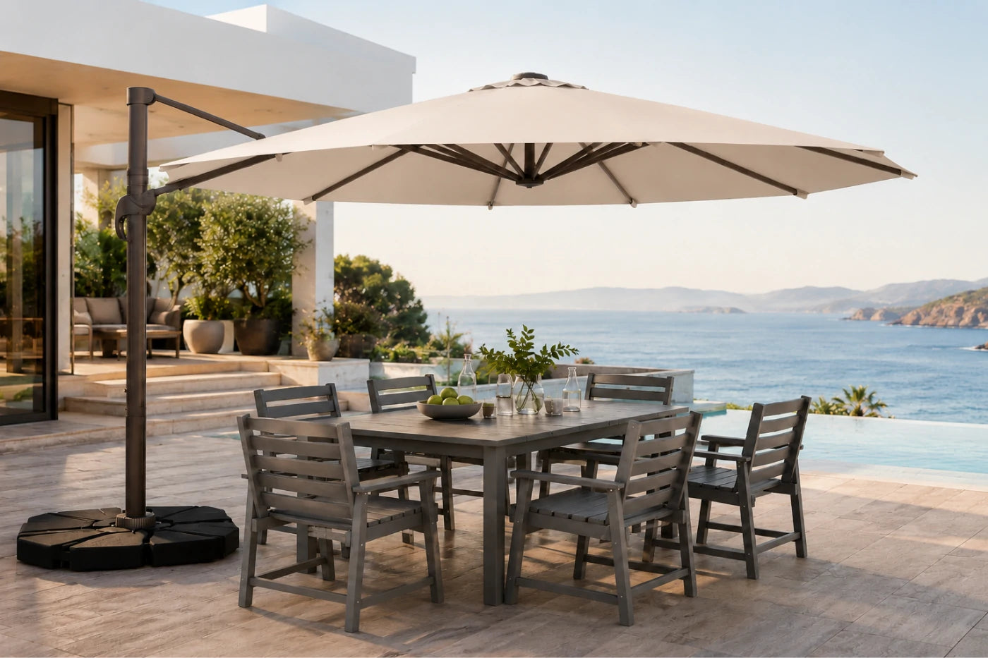 An outdoor patio dining set with a rectangular gray table and six matching slat-back chairs under a large beige patio umbrella, set on a stone terrace overlooking the sea.