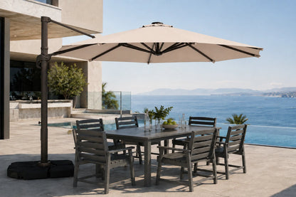 A large outdoor patio umbrella with a dark metal cantilever arm and black base, shading a rectangular gray dining table and six matching slatted chairs on a sunlit terrace overlooking a blue ocean and pool.