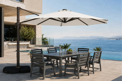 A large outdoor patio umbrella with a dark metal cantilever arm and black base, shading a rectangular gray dining table and six matching slatted chairs on a sunlit terrace overlooking a blue ocean and pool.