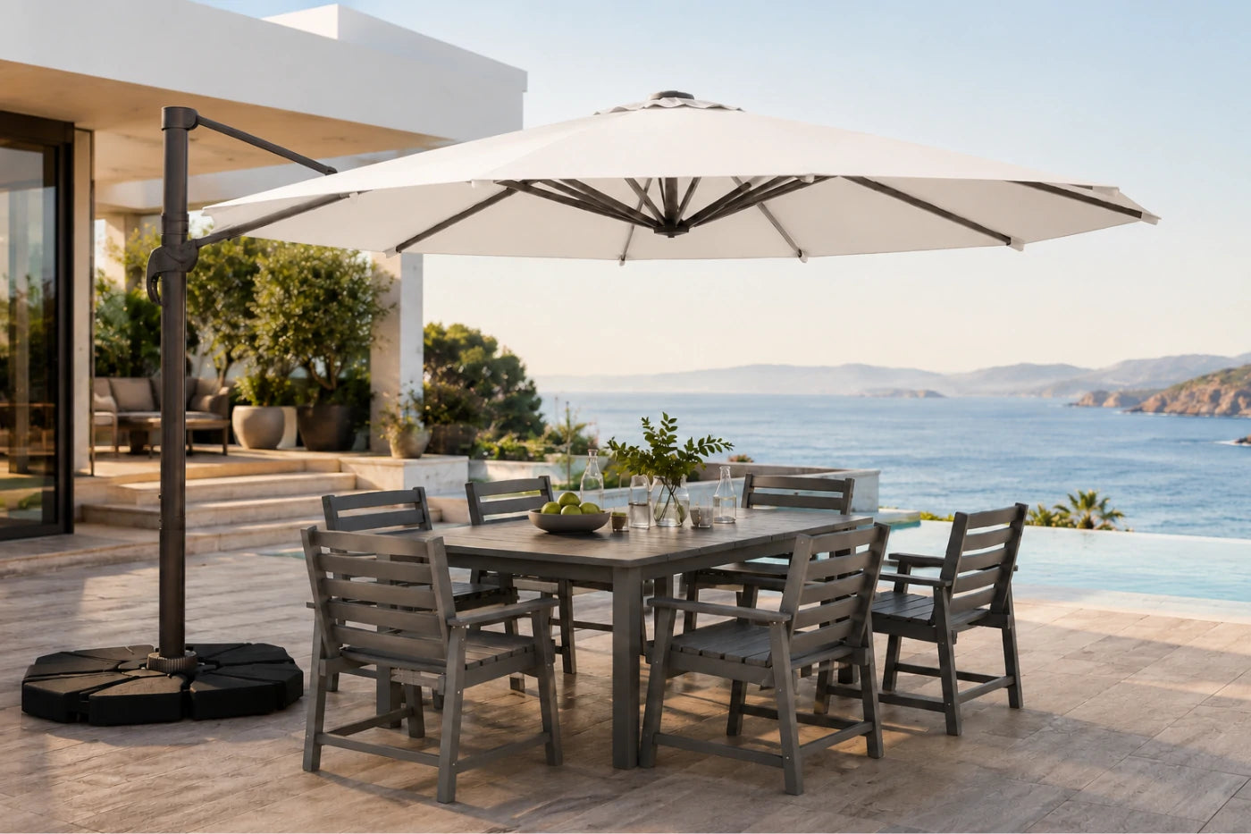 A large patio umbrella with a black cantilever pole and weight base, shading a gray outdoor dining table and six chairs on a stone terrace overlooking a blue ocean and sea walls.