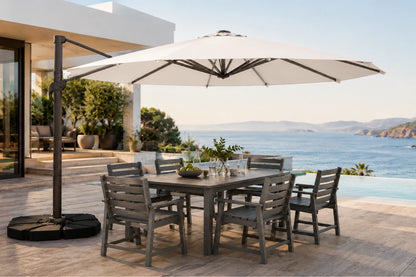 A large patio umbrella with a black cantilever pole and weight base, shading a gray outdoor dining table and six chairs on a stone terrace overlooking a blue ocean and sea walls.