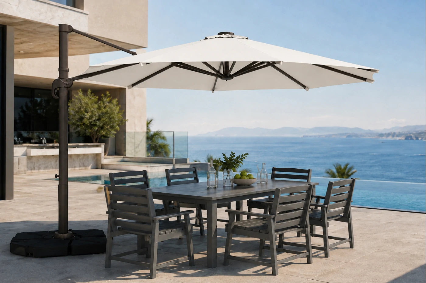 A large outdoor patio umbrella with a dark metal cantilever arm and black base, shading a rectangular gray dining table and six matching slatted chairs on a sunlit terrace overlooking a blue ocean and pool.