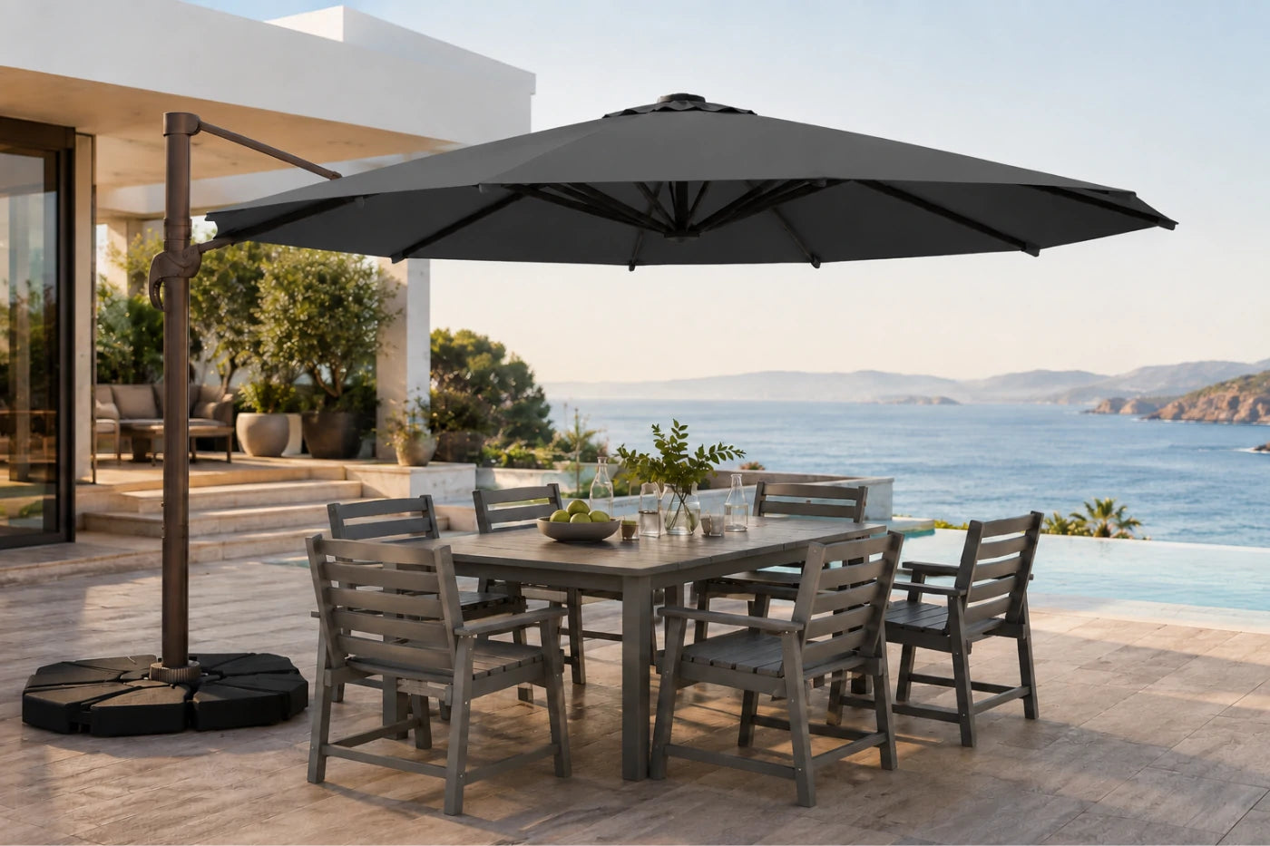 A large dark gray cantilever patio umbrella with a square base is shown outdoors on a stone terrace beside a dining table and chairs, with a pool and ocean view in the background.