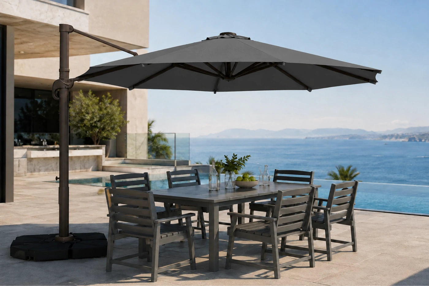 A large outdoor patio umbrella with a dark metal cantilever arm and black base, shading a rectangular gray dining table and six matching slatted chairs on a sunlit terrace overlooking a blue ocean and pool.
