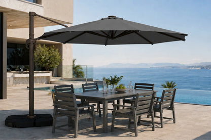 A large outdoor patio umbrella with a dark metal cantilever arm and black base, shading a rectangular gray dining table and six matching slatted chairs on a sunlit terrace overlooking a blue ocean and pool.