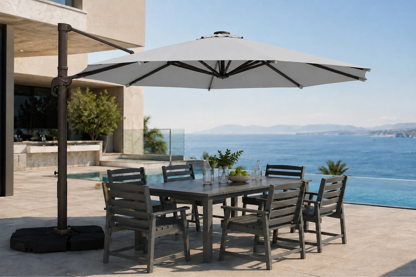 A large outdoor patio umbrella with a dark metal cantilever pole and base is shown on a sunlit terrace beside a dining table and chairs, with an ocean view in the background.