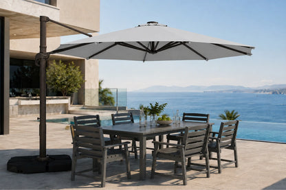 A large outdoor patio umbrella with a dark metal cantilever pole and base is shown on a sunlit terrace beside a dining table and chairs, with an ocean view in the background.