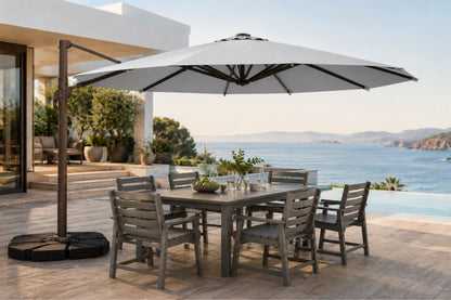 A large outdoor patio dining set with a rectangular gray table and six matching slatted chairs under a wide white cantilever umbrella, set on a stone terrace overlooking a blue ocean and sea view.