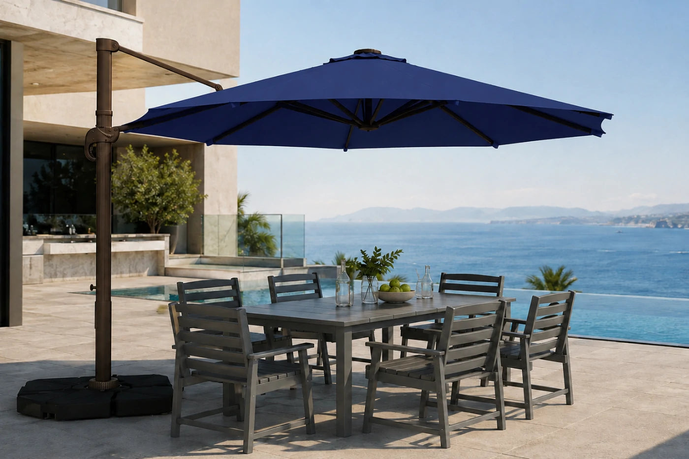 A navy blue cantilever patio umbrella with a dark metal pole and black weight-bearing base, shading a gray wooden outdoor dining table and six matching slatted chairs on a patio. The background features a modern building, a swimming pool, and a scenic ocean view under a clear blue sky.
