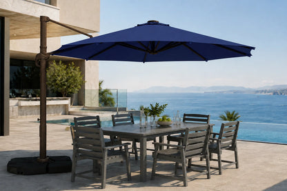 A large outdoor patio umbrella with a dark metal cantilever arm and black base, shading a rectangular gray dining table and six matching slatted chairs on a sunlit terrace overlooking a blue ocean and pool.