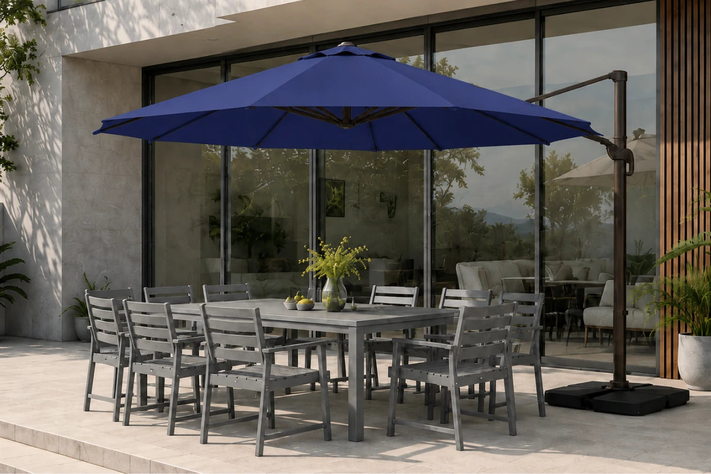 A large navy blue cantilever patio umbrella with a black metal frame and base, shading a gray outdoor dining table and chairs on a stone patio in front of a modern glass building.