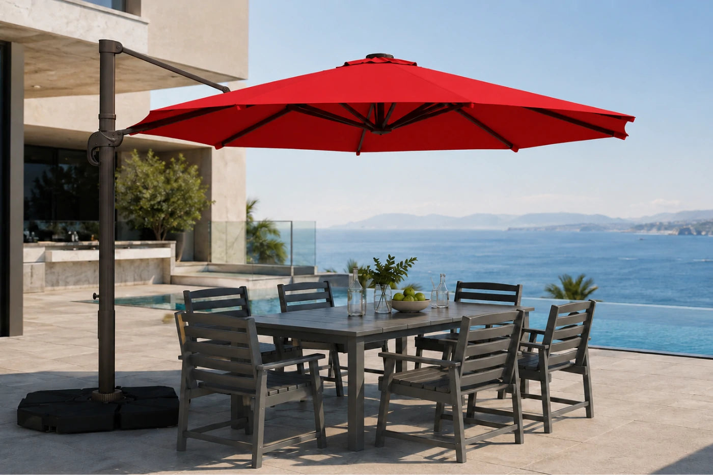 A large red patio umbrella with a dark metal pole and weighted base, shading a rectangular outdoor dining table and six gray slat chairs on a patio overlooking a blue ocean and distant mountains.